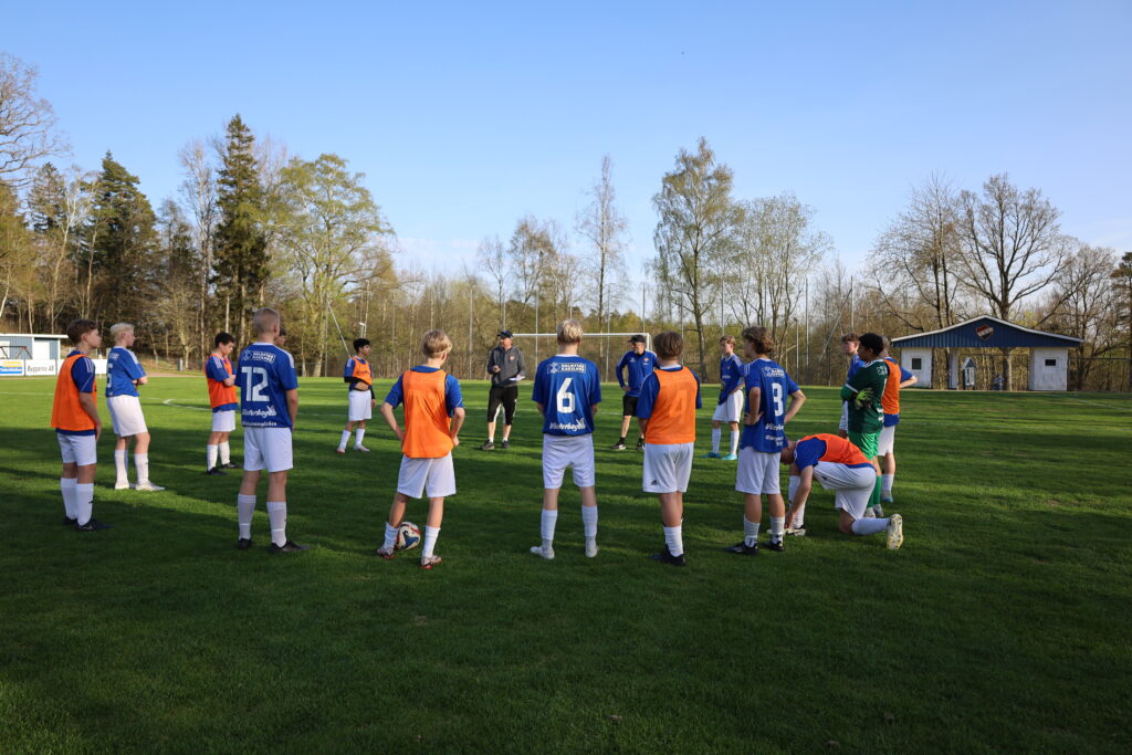 Laget står i en ring på fotbollsplanen. Tränarna står också i ringen och pratar med spelarna.