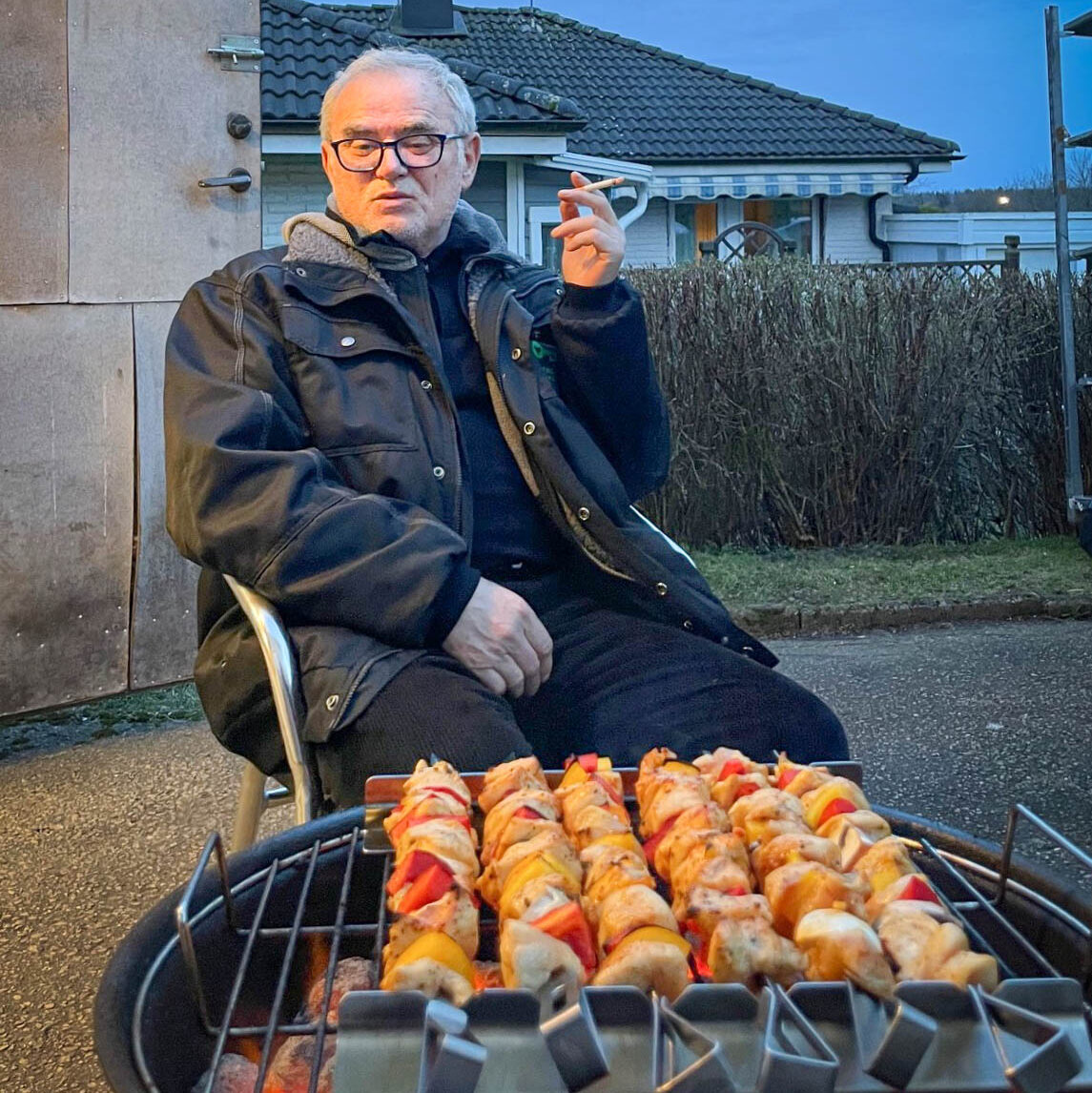 Senad sitter framför grillen med en cigarett i handen. På grillen ligger kyckling- och paprikaspett.